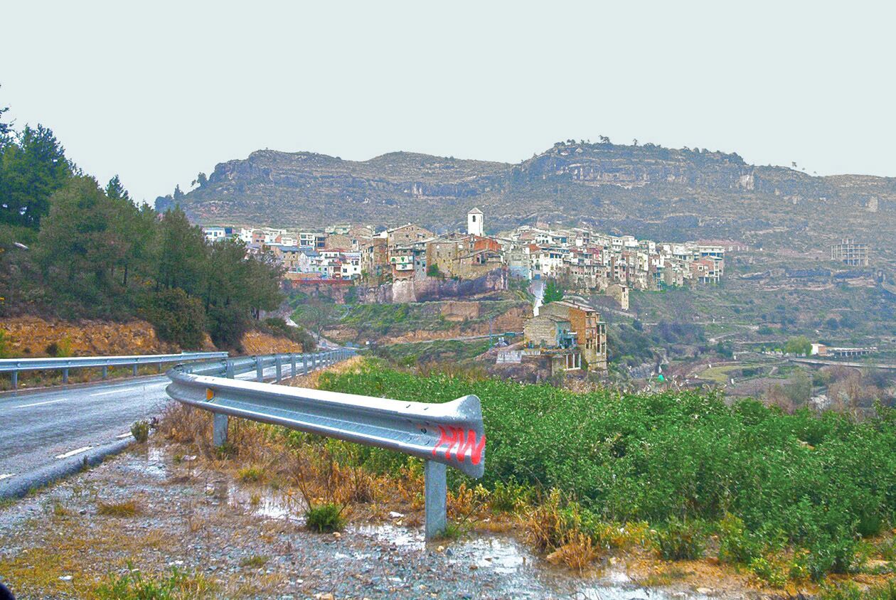 A coastal road with a guardrail, a village with colorful buildings, and a hill in the background.