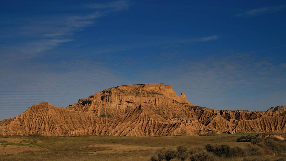 A large, rugged butte with a flat top and steep sides, surrounded by a flat, grassy plain under a clear blue sky.
