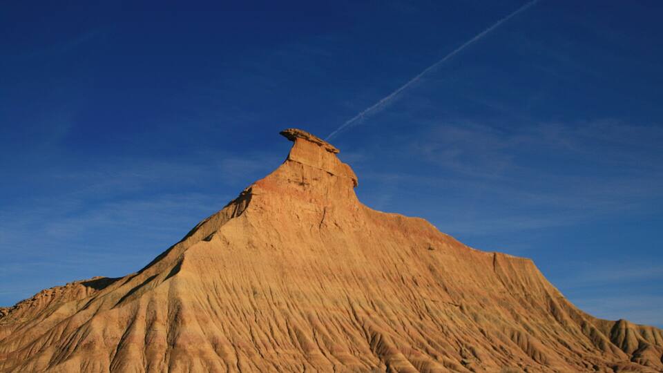 A large, steep, reddish-brown rock formation with a flat top and a visible trail or path leading up to it.