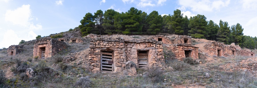 Ruinas de piedra antiguas con múltiples puertas y ventanas, rodeadas de vegetación escasa y pinos.