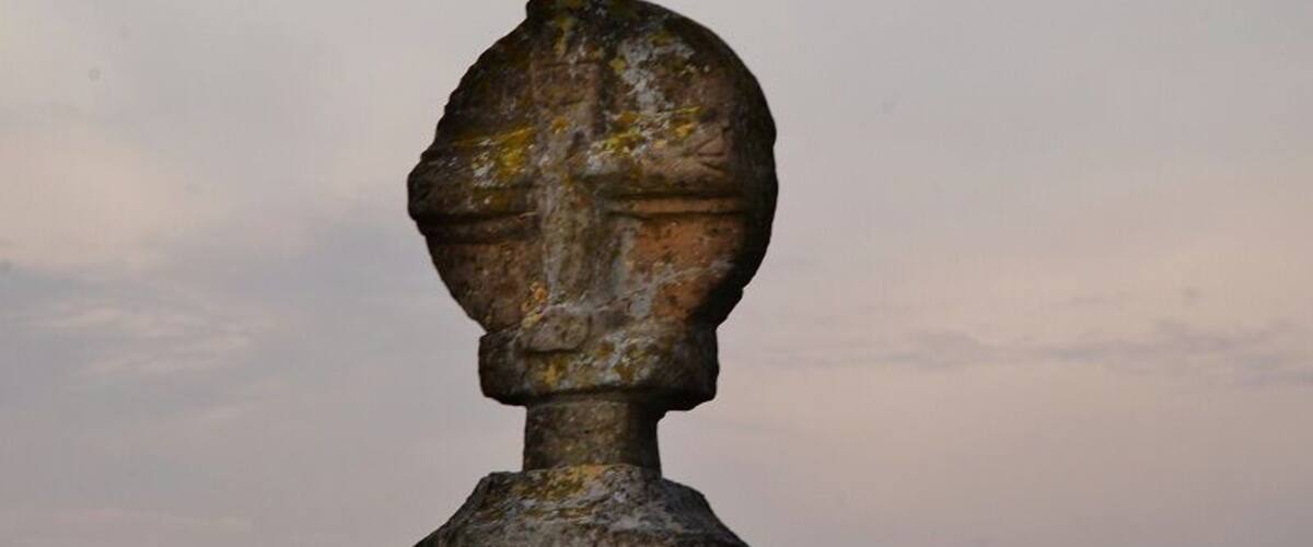 A weathered head sculpture on a pedestal against a cloudy sky.