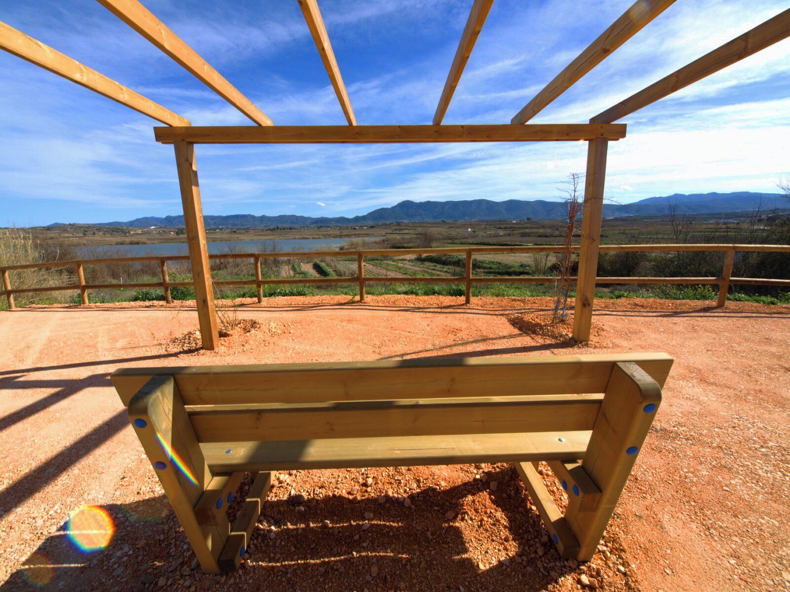 A wooden bench in a dirt field with a metal railing, overlooking a scenic landscape with mountains and a body of water.