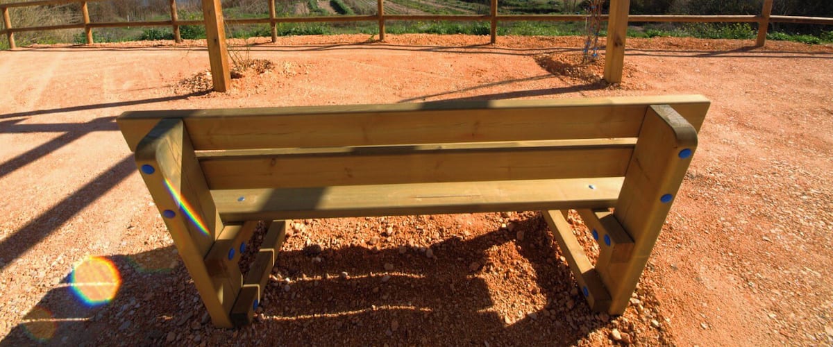 A wooden bench in a dirt field with a metal railing, overlooking a scenic landscape with mountains and a body of water.