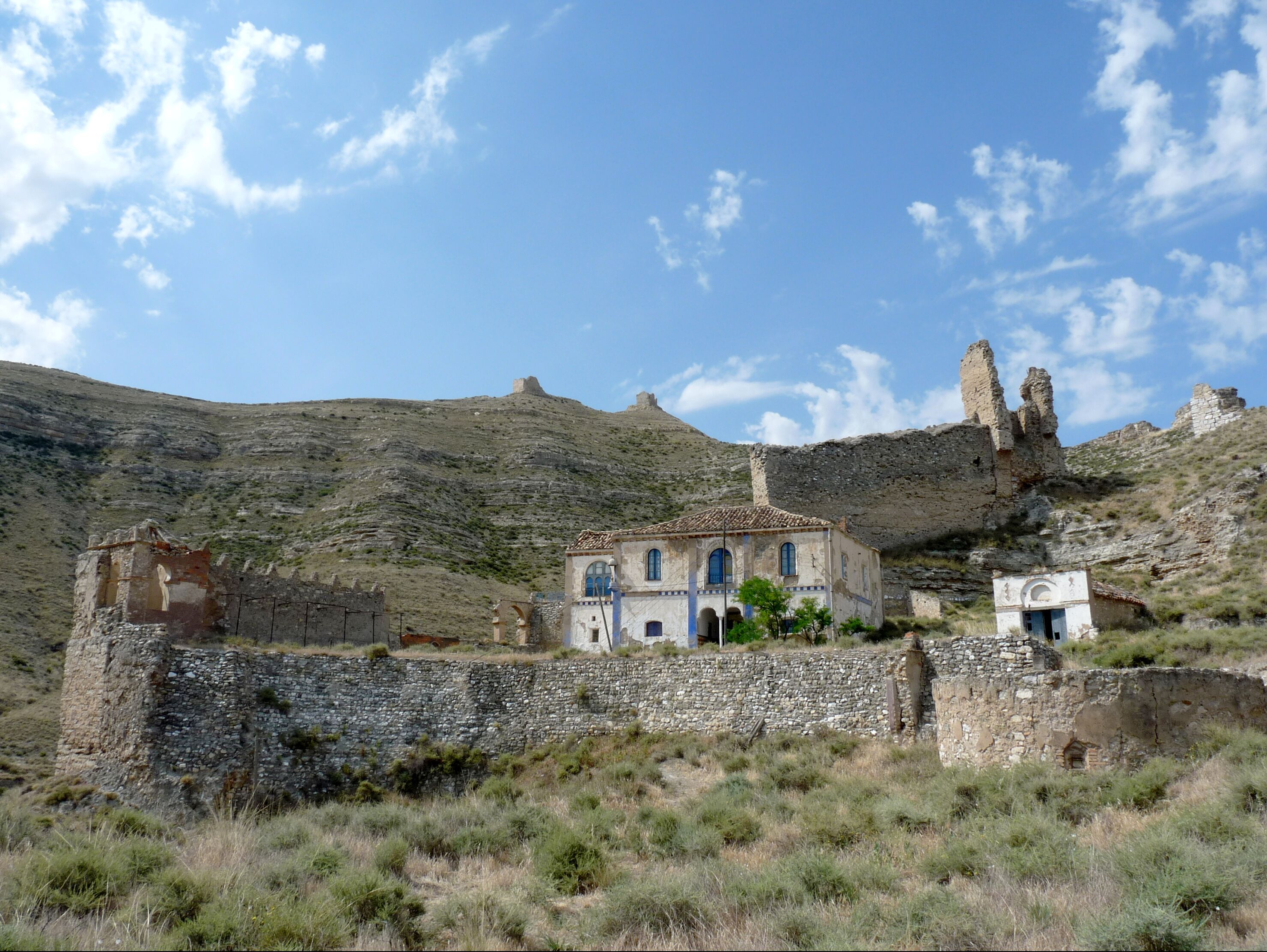 A historical building with blue doors and windows, surrounded by stone walls and ruins on a hillside.