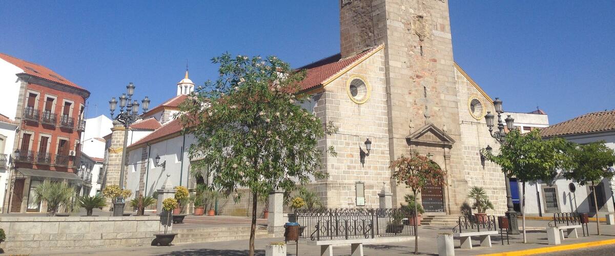 A historic church with a tall bell tower and a clock, surrounded by traditional buildings and a clear blue sky.