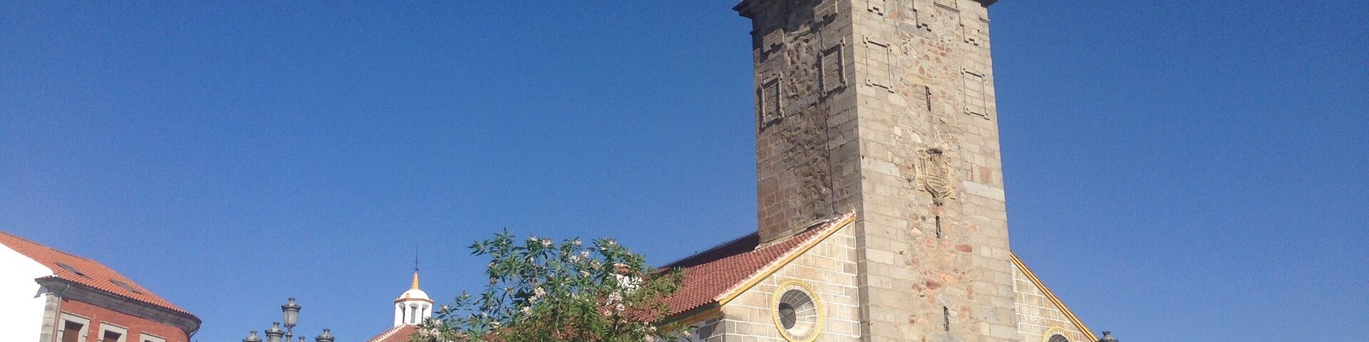 A historic church with a tall bell tower and a clock, surrounded by traditional buildings and a clear blue sky.