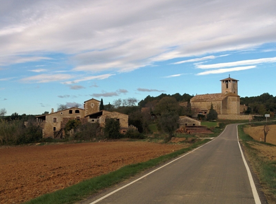 A rural road curving through fields with stone buildings and a church with a bell tower.