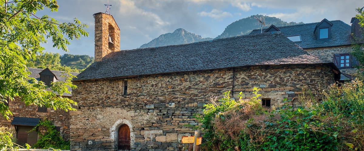 A stone building with a wooden door, surrounded by greenery and a mountain in the background.