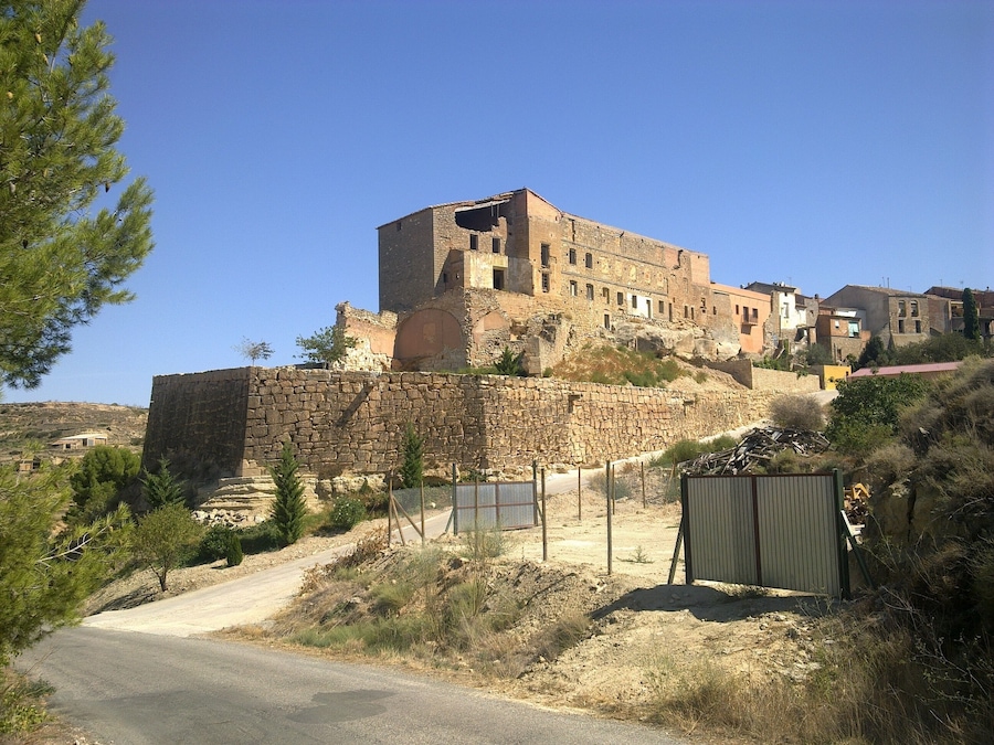 Uma fortaleza de pedra histórica com uma grande torre central, cercada por uma muralha de pedra e uma estrada de terra.