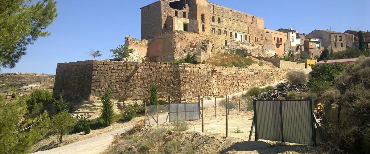 A historic stone fortress with a large central tower, surrounded by a stone wall and a dirt road.