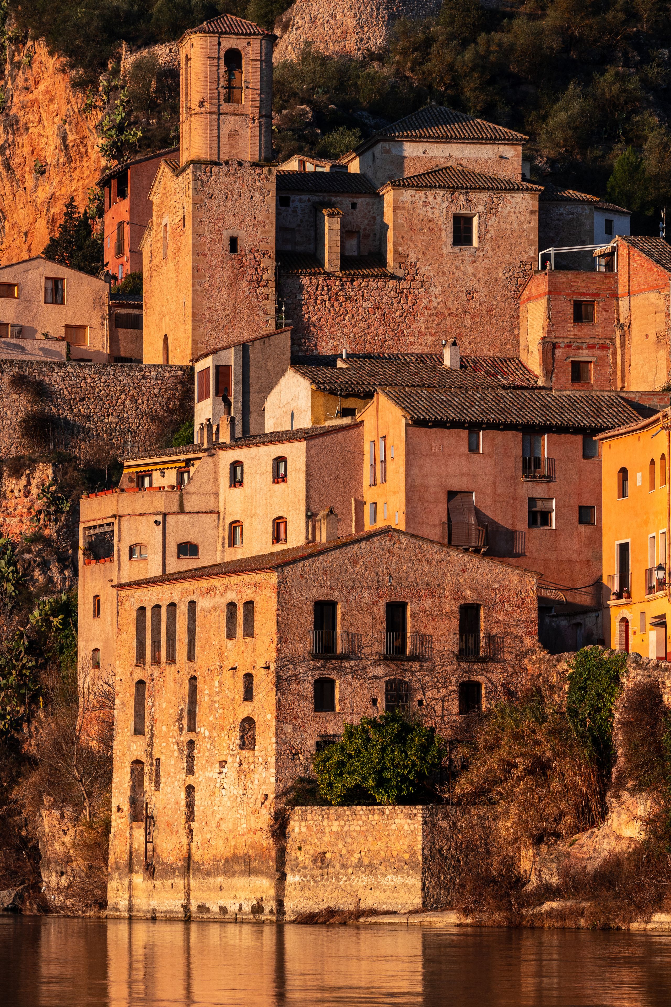 A historic town with terracotta-roofed buildings, a stone wall, and a river.