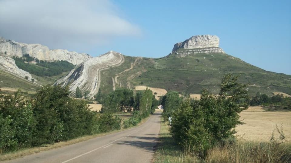 A road leading to a large white rock formation with a forested hill in the background.