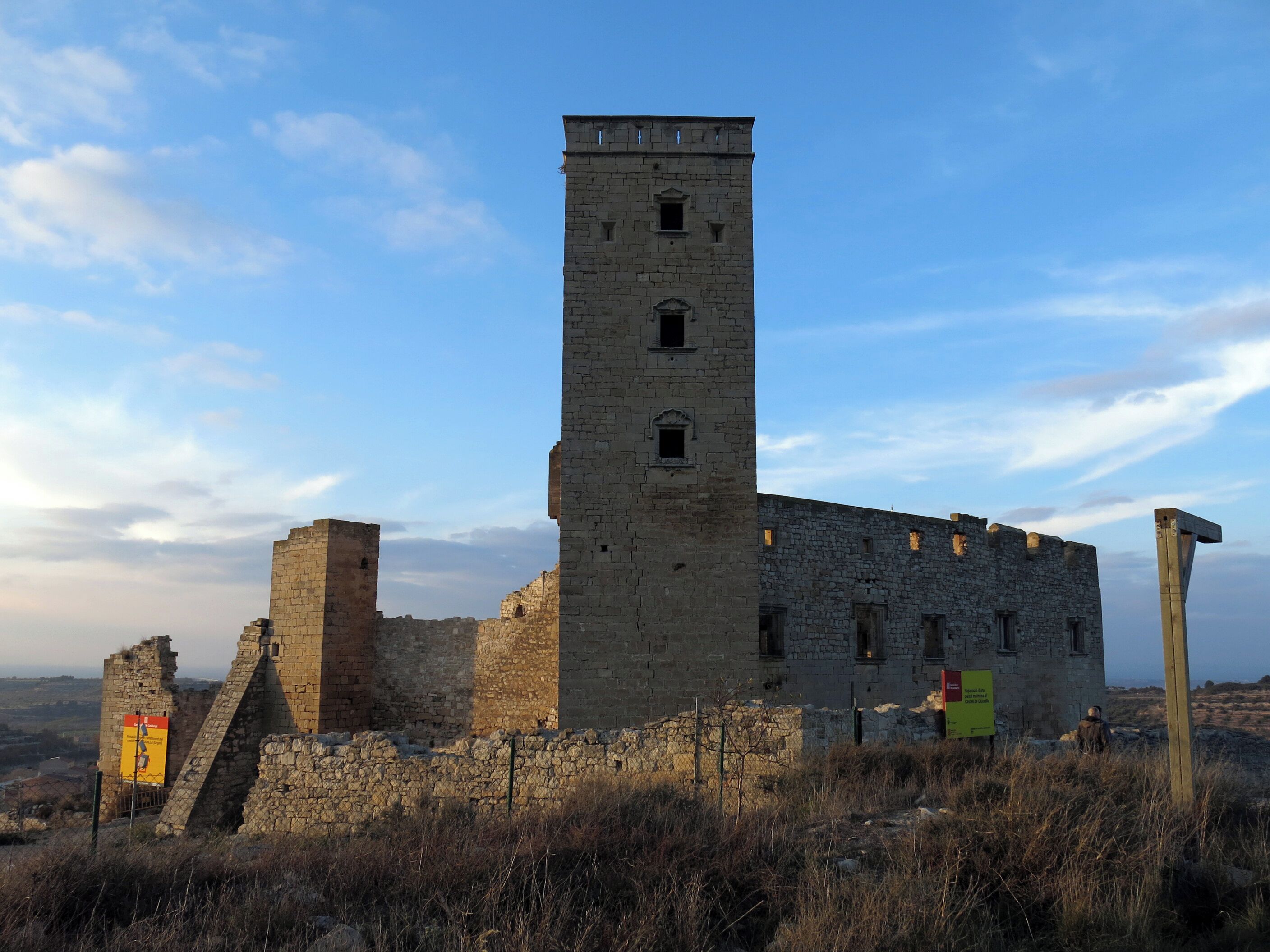 A medieval stone tower with a rectangular base and a smaller tower on top, surrounded by dry grass and a clear sky.