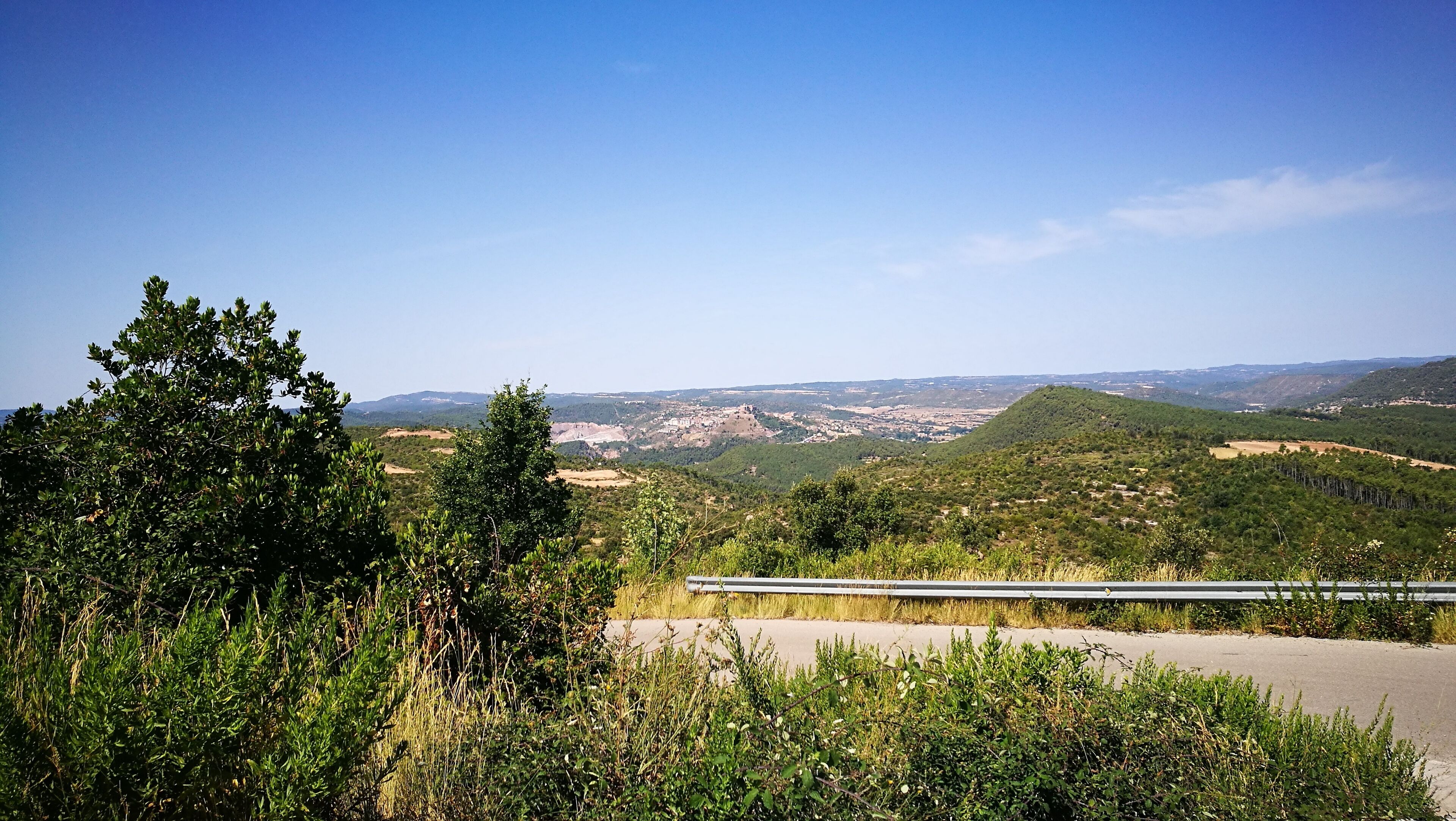 A road with a guardrail, surrounded by dense greenery and hills.