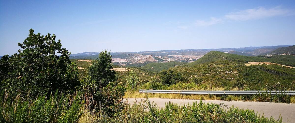A road with a guardrail, surrounded by dense greenery and hills.