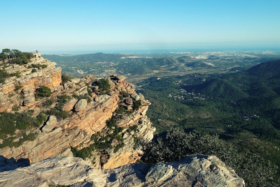 A rocky cliff overlooking a valley with scattered buildings and greenery.