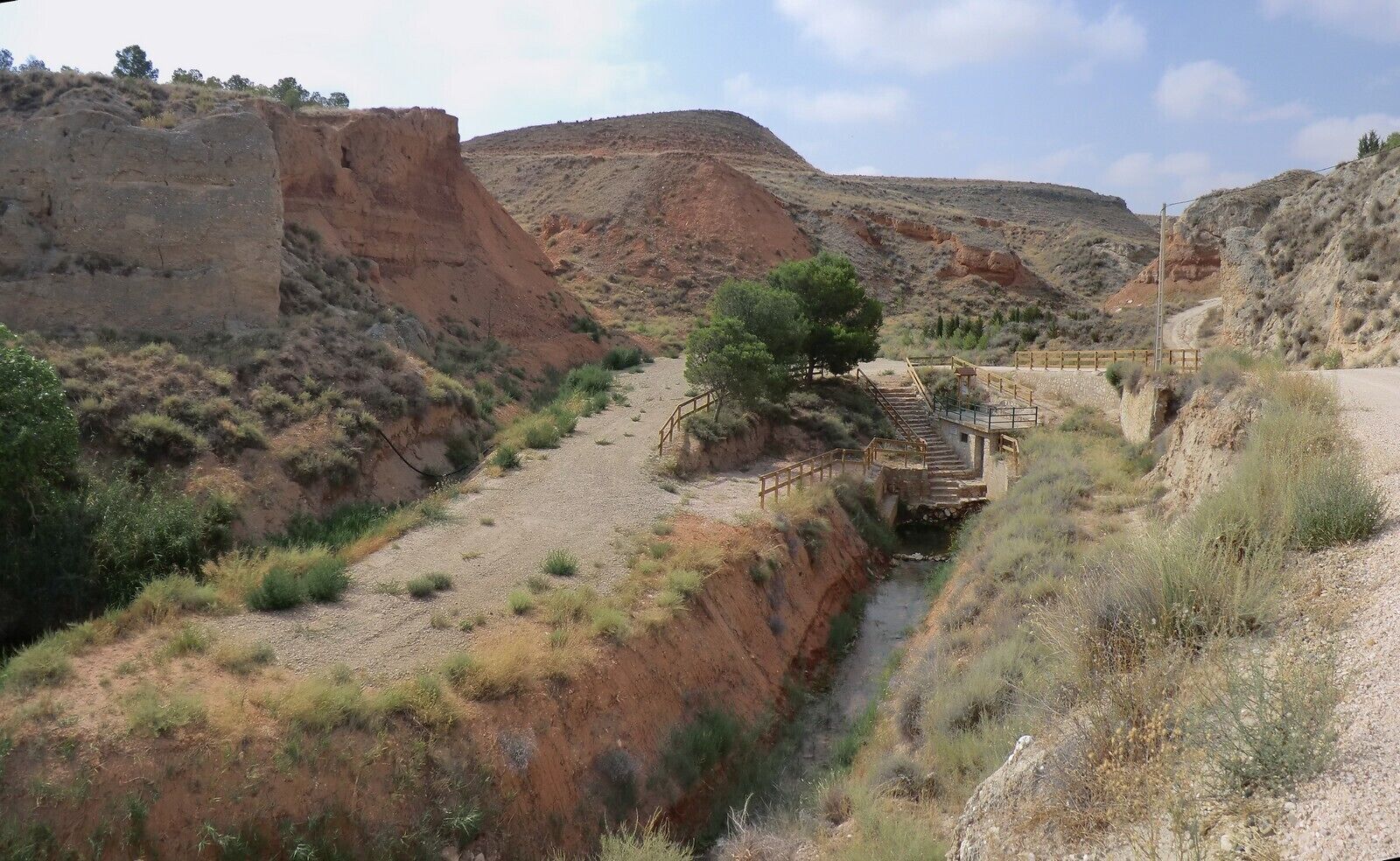 A dry riverbed with a wooden bridge and steps leading over it, surrounded by reddish-brown hills and sparse vegetation.