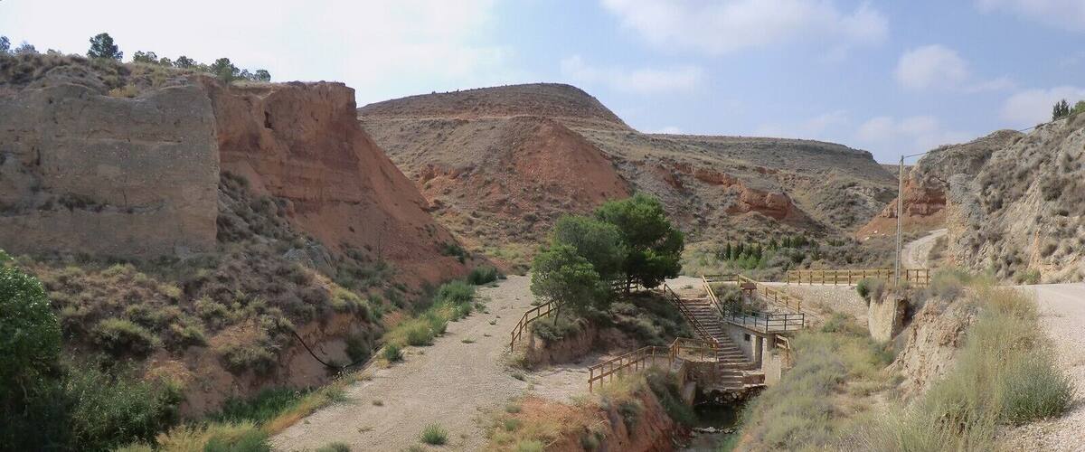 A dry riverbed with a wooden bridge and steps leading over it, surrounded by reddish-brown hills and sparse vegetation.