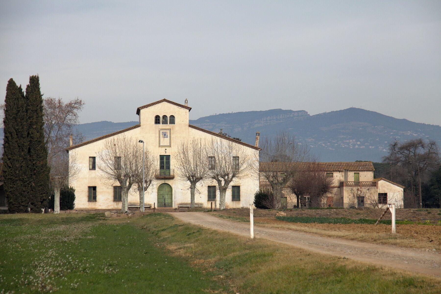 A large, single-story building with a tiled roof, surrounded by trees and a dirt path.