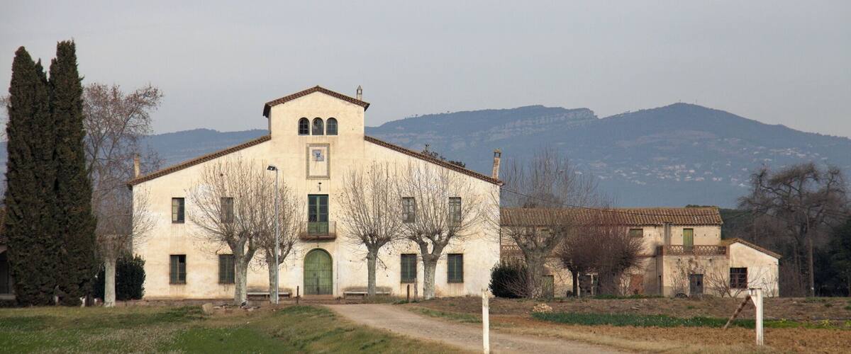 A large, single-story building with a tiled roof, surrounded by trees and a dirt path.