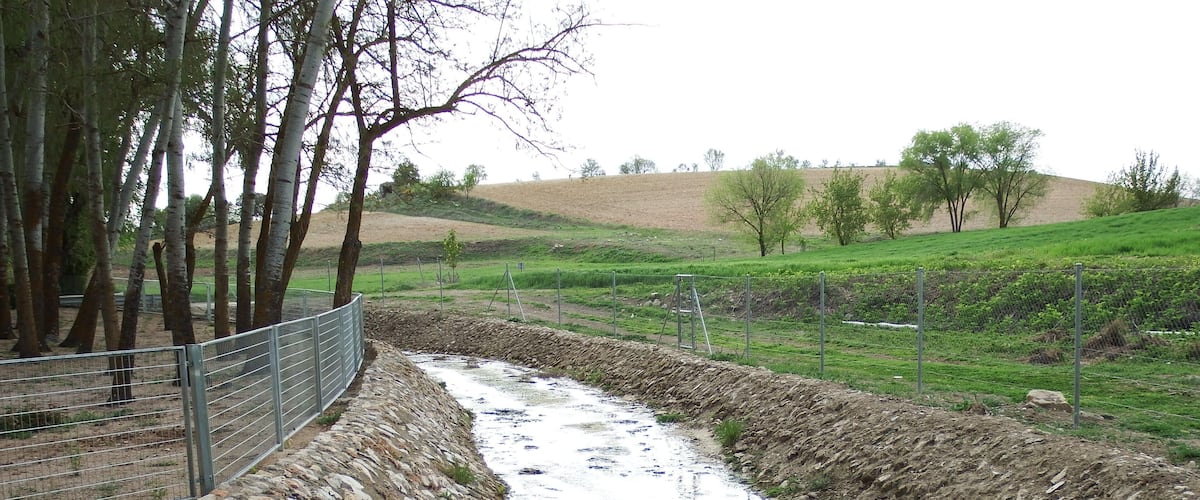 A narrow stream flowing through a stone-lined channel, surrounded by trees and grassy fields.