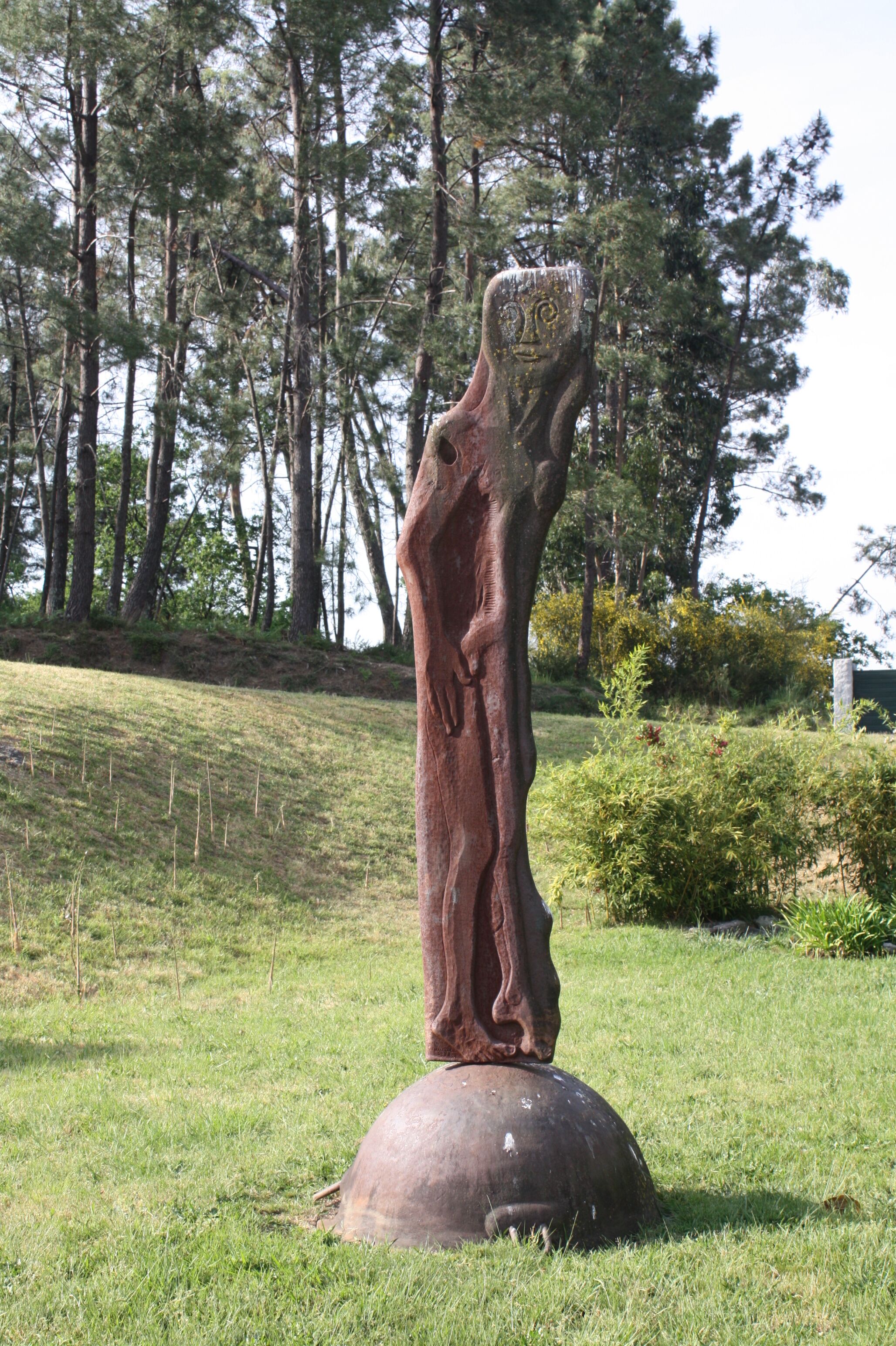 A sculpture of a human figure with a tree trunk for a body, standing on a large stone base, surrounded by grass and trees.
