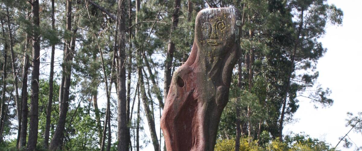 A sculpture of a human figure with a tree trunk for a body, standing on a large stone base, surrounded by grass and trees.