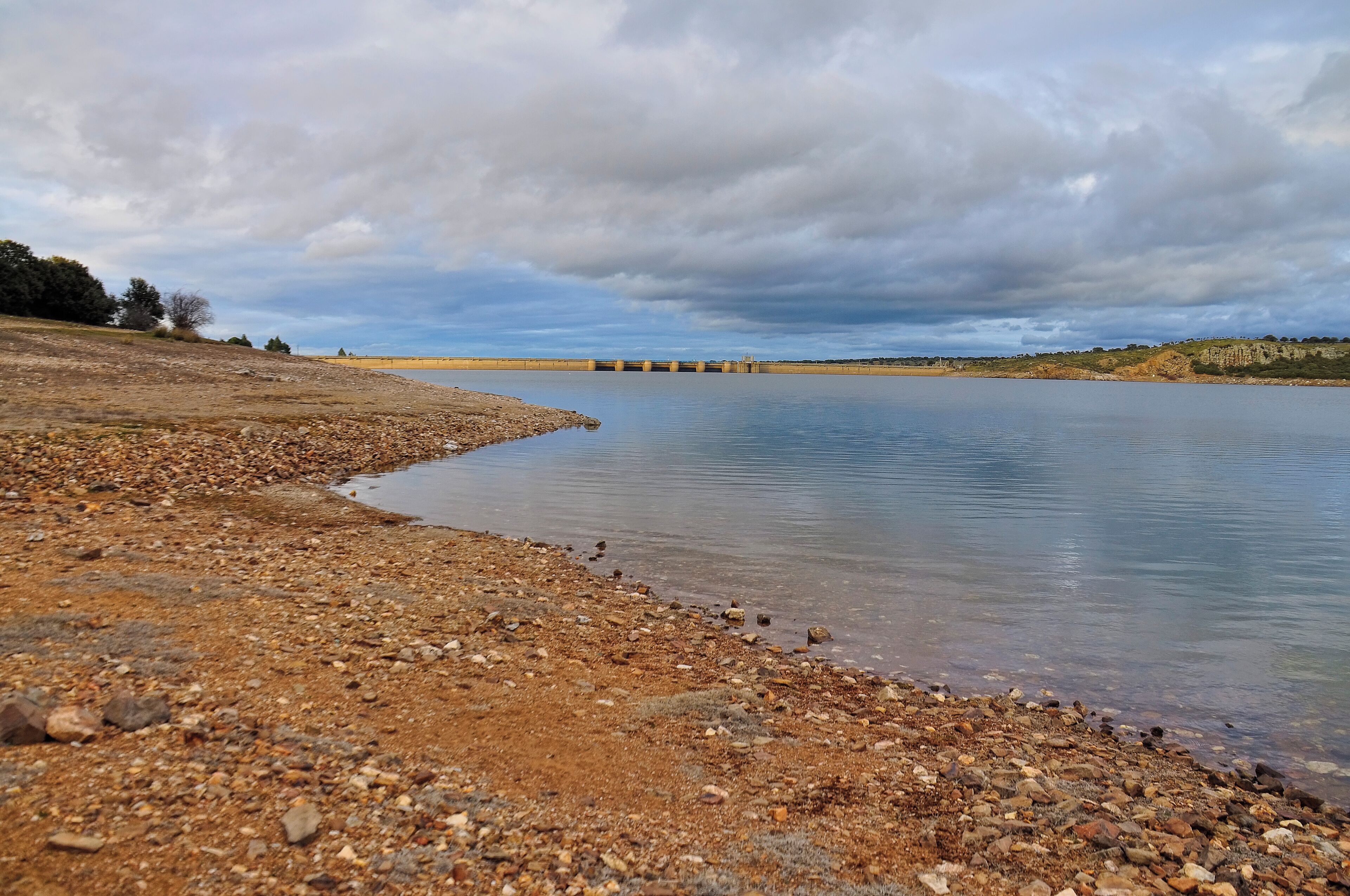 A rocky shoreline with a calm lake, a distant dam, and overcast sky.