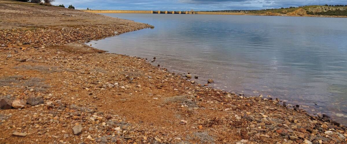 A rocky shoreline with a calm lake, a distant dam, and overcast sky.