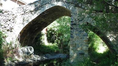 Un vieux pont de pierre en arc de cercle enjambant un ruisseau, entouré d’une végétation luxuriante et d’arbres.