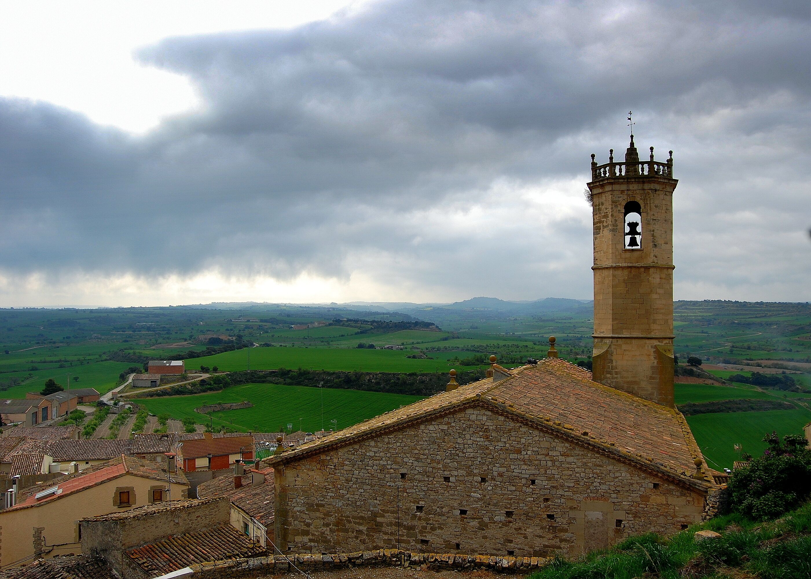 A stone church with a bell tower, surrounded by green fields and rolling hills.