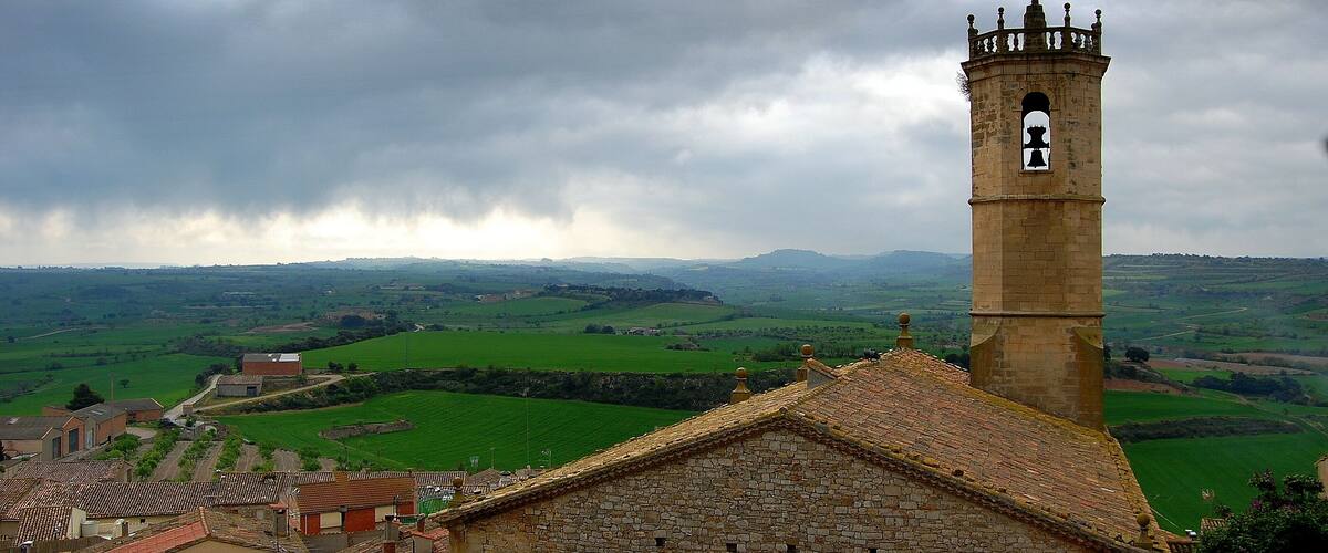 A stone church with a bell tower, surrounded by green fields and rolling hills.