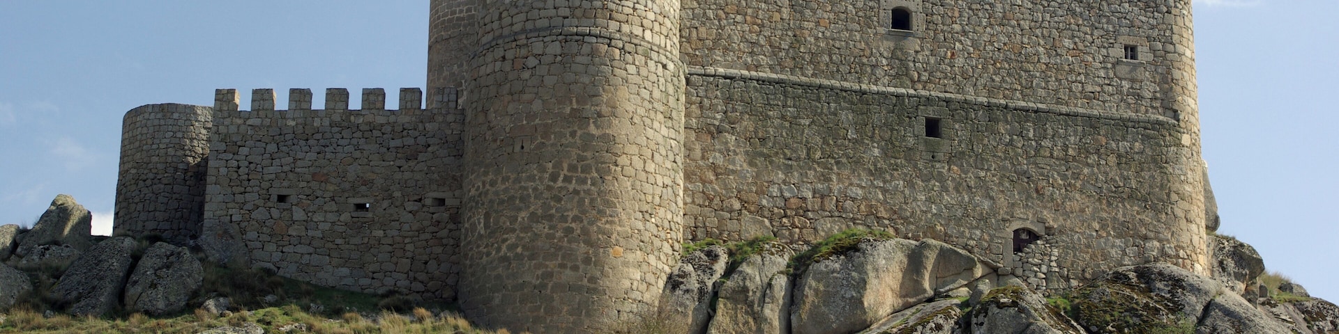 Un castillo de piedra medieval con múltiples torres y almenas, asentado sobre una colina rocosa con vegetación escasa.