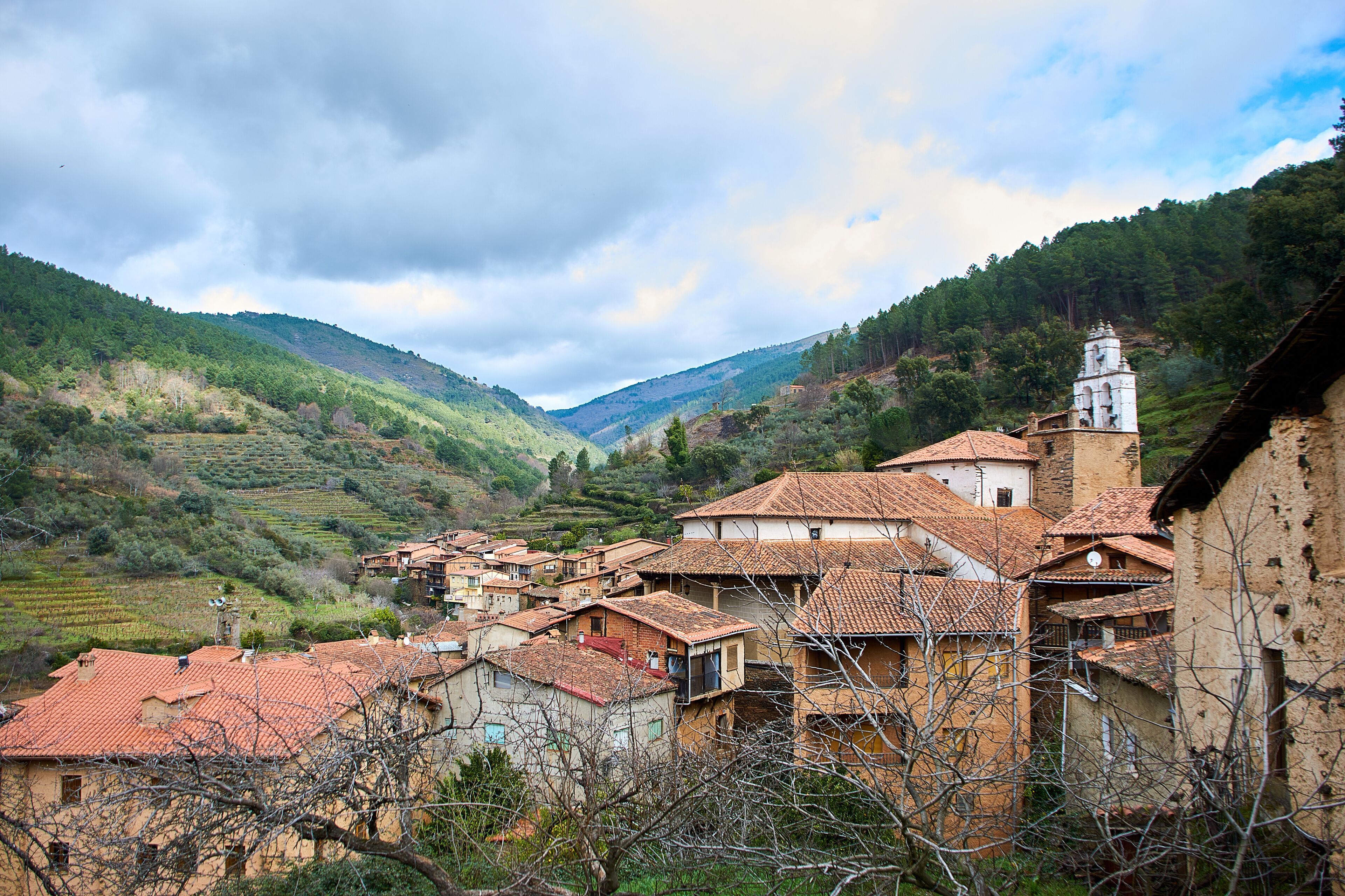 A village with terracotta-roofed houses nestled in a valley, surrounded by lush green hills and a prominent church tower.