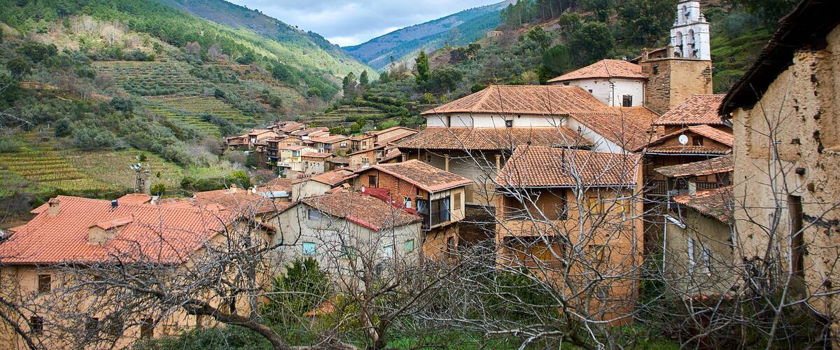 A village with terracotta-roofed houses nestled in a valley, surrounded by lush green hills and a prominent church tower.