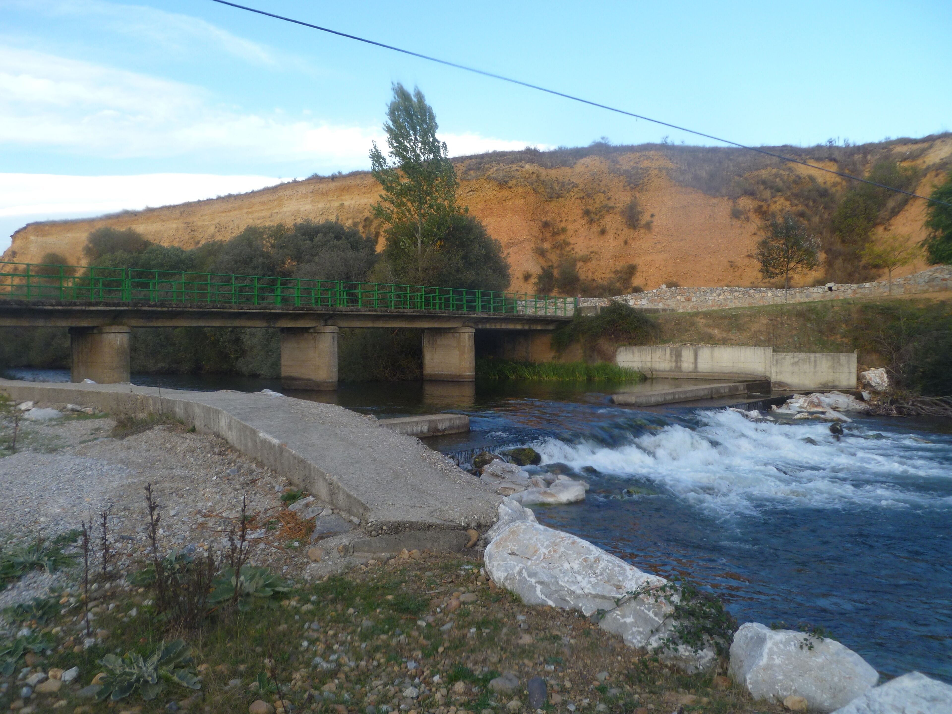 A concrete bridge over a fast-flowing river with a rocky riverbank and a hillside in the background.