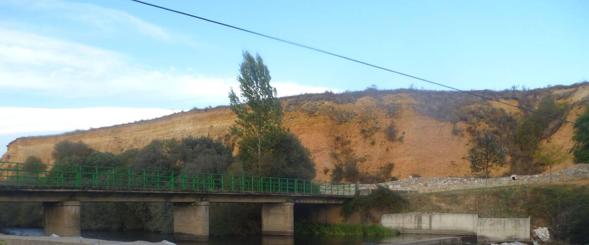 A concrete bridge over a fast-flowing river with a rocky riverbank and a hillside in the background.