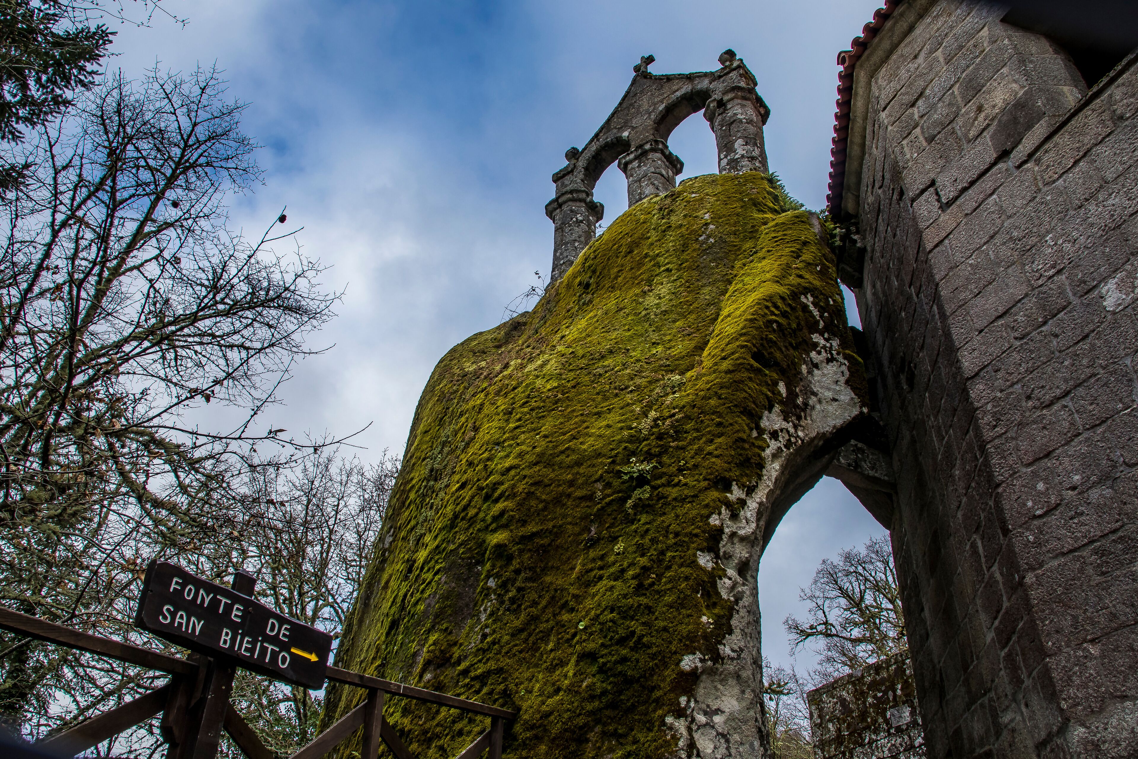 A moss-covered stone structure with an archway and a sign indicating 'Fonte de San Bieito'.