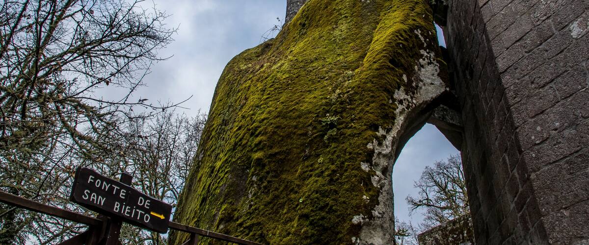 A moss-covered stone structure with an archway and a sign indicating 'Fonte de San Bieito'.