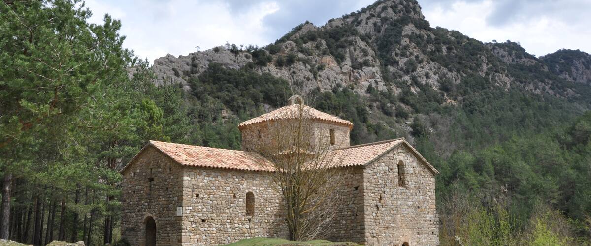 A stone church with a tiled roof surrounded by a forest and mountains.