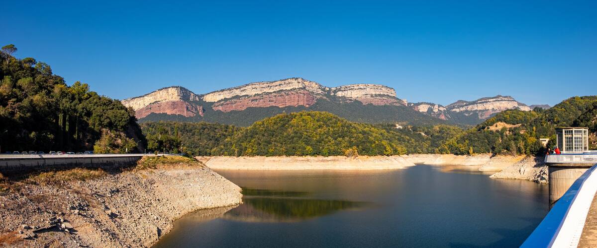 A dam with a bridge, calm water, and mountains in the background.