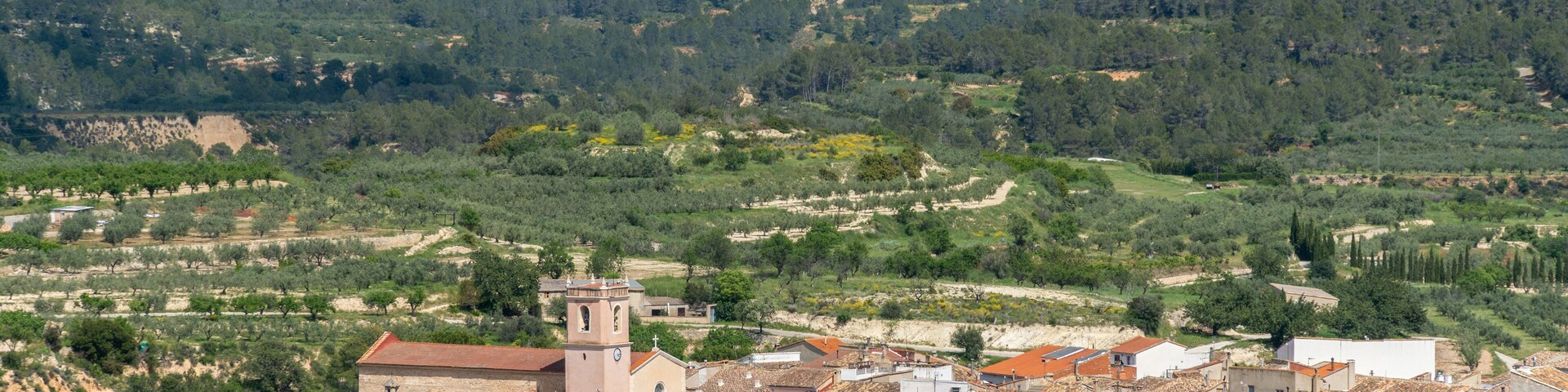 Uma vila com edifícios de telhados em terracota, uma igreja imponente com uma torre de sino, e ao fundo colinas e florestas verdes.