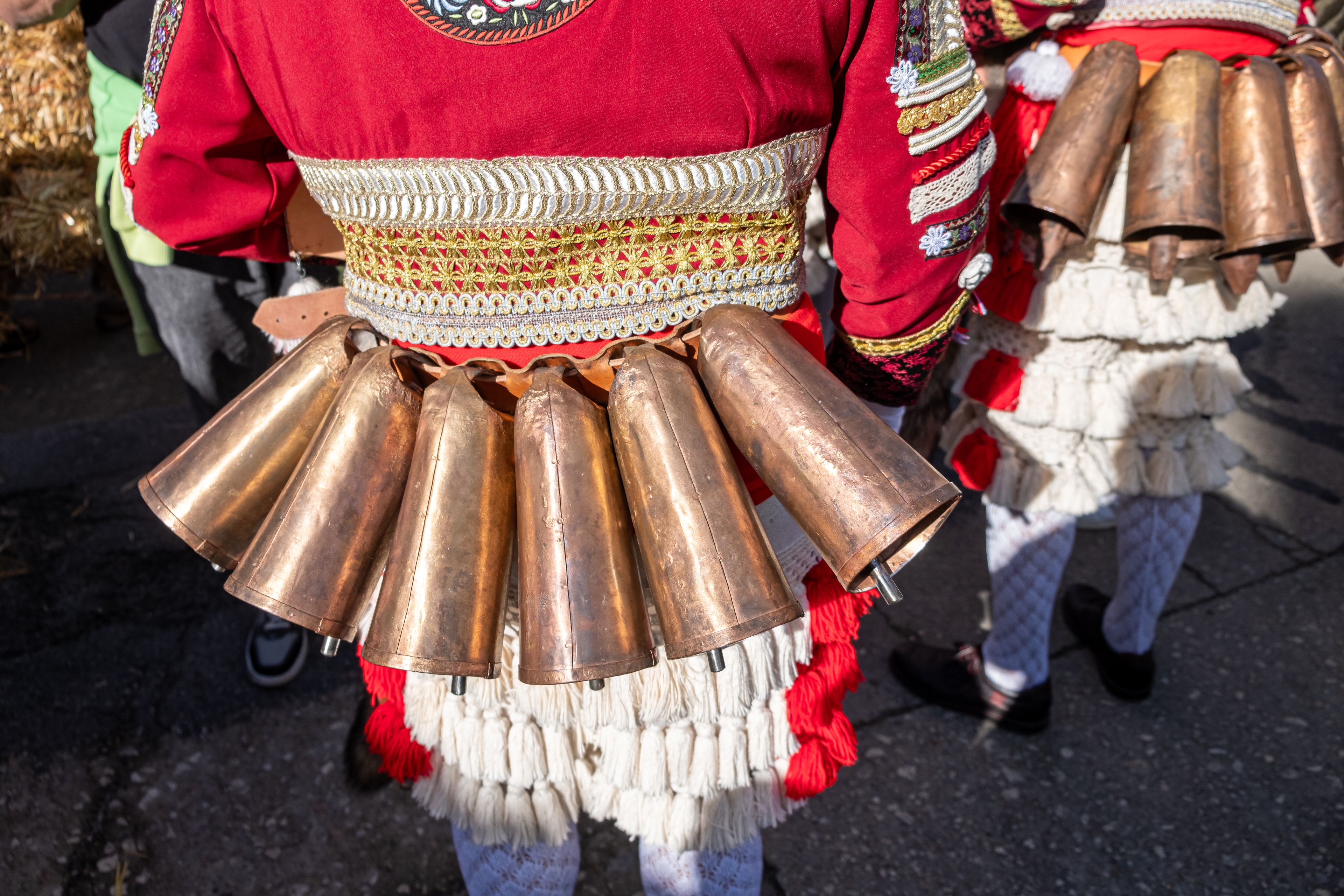 A person wearing a red garment with a decorative belt and multiple copper bells.