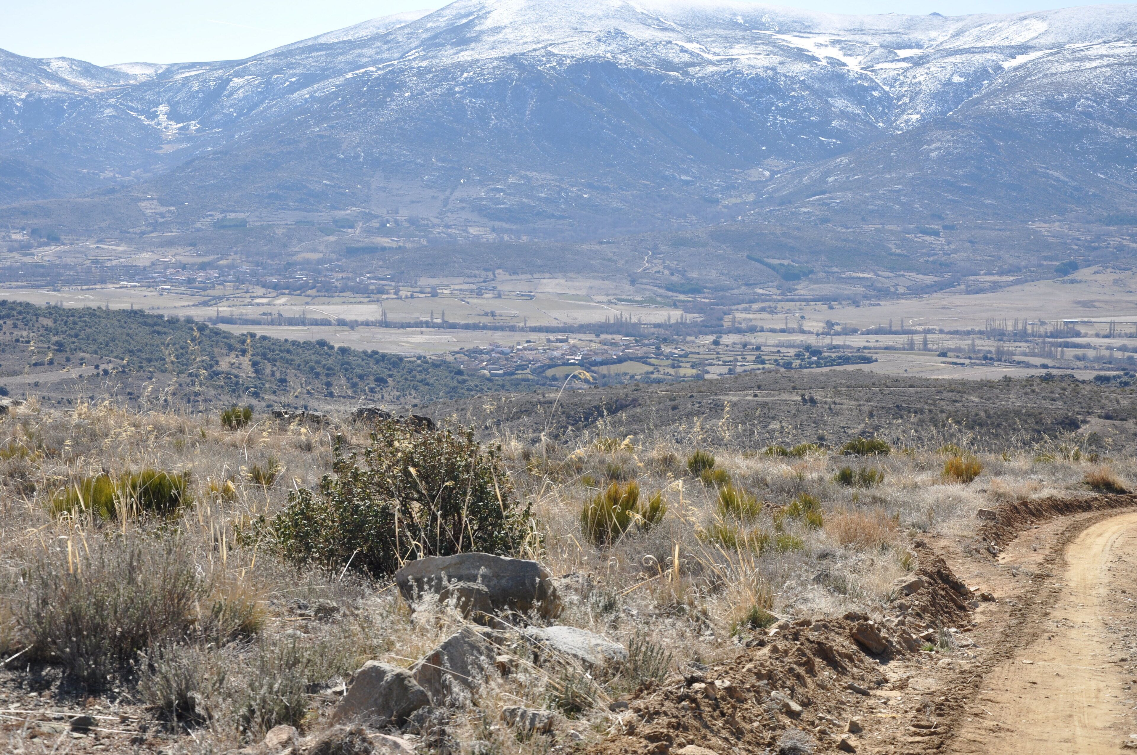 A dirt road winding through a dry, rocky landscape with sparse vegetation and a snow-capped mountain in the background.