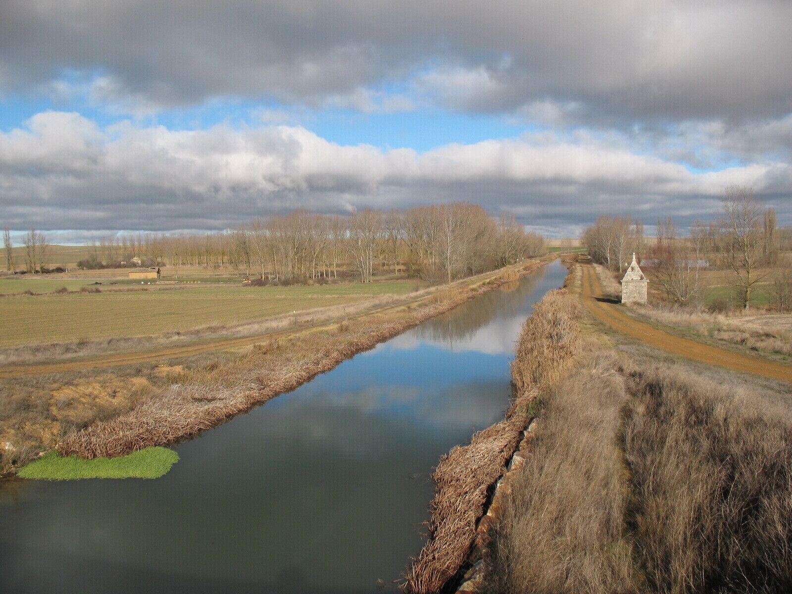 A river with a stone wall on one side and a dirt road on the other, surrounded by fields and trees.