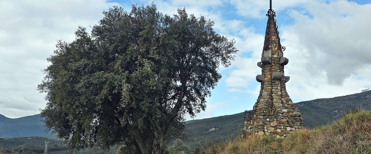 A stone cross monument, a large tree, and a rural landscape with houses in the background.