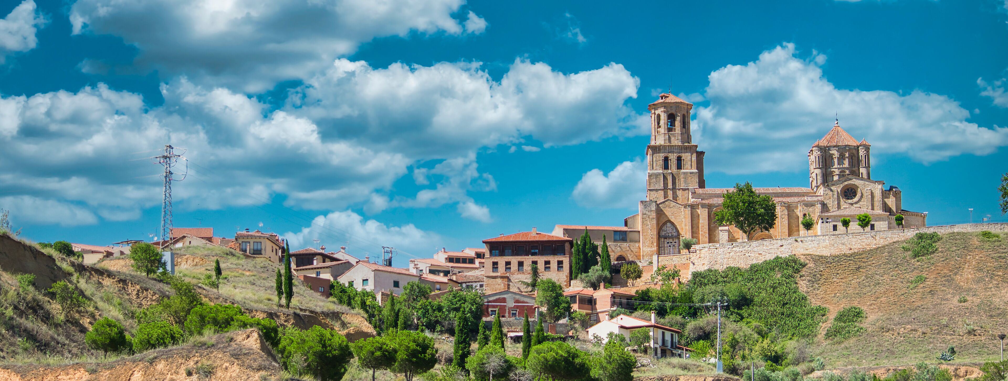 A historic church with a bell tower and a stone wall, surrounded by houses on a hillside under a blue sky.