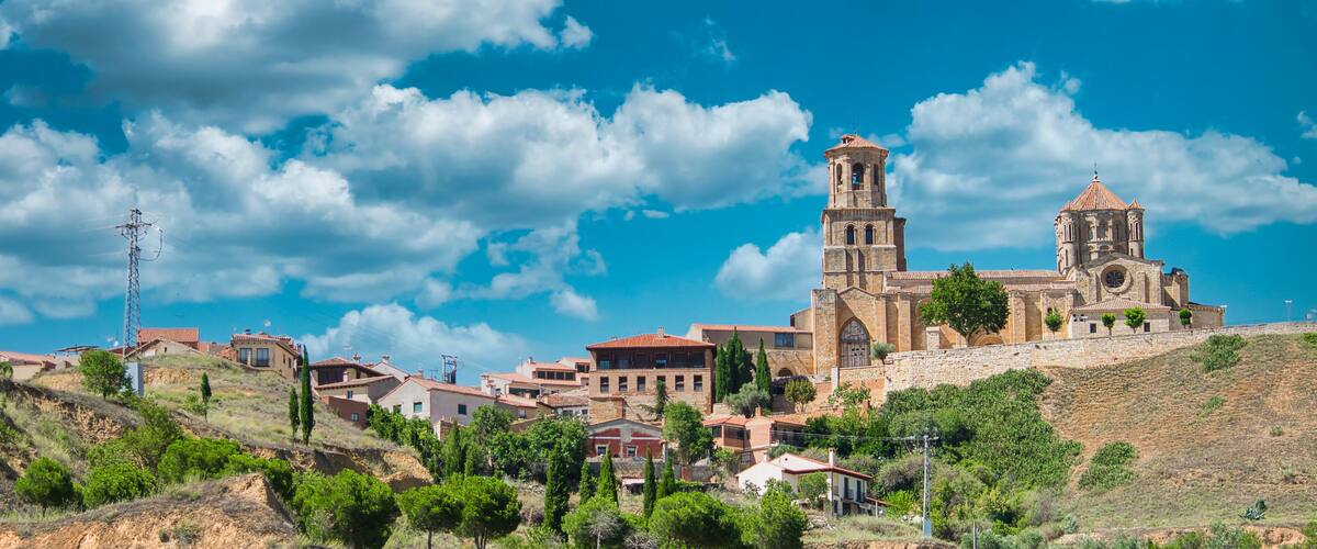 A historic church with a bell tower and a stone wall, surrounded by houses on a hillside under a blue sky.