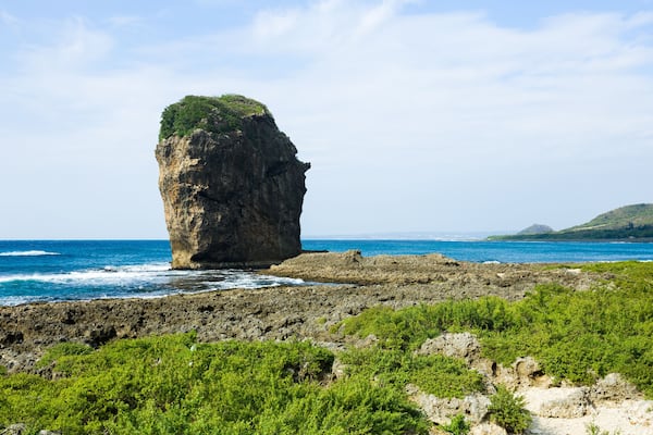 A prominent rock formation with a green top, surrounded by ocean and coastal vegetation.