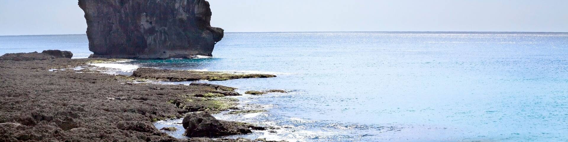 A rocky coastline with a prominent rock formation surrounded by clear blue waters and a partly cloudy sky.